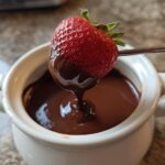 A fresh strawberry being dipped into a small white pot of smooth, melted chocolate fondue.