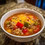 A close-up of a white bowl filled with savory stuffed pepper soup, featuring ground meat, rice, colorful peppers, and melted cheddar cheese.