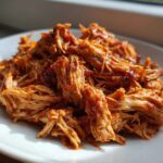 Close-up of tender, saucy pulled chicken piled high on a white plate, ready to serve.