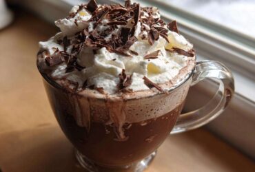 A clear glass mug filled with rich boozy hot chocolate, topped high with whipped cream and dark chocolate shavings.