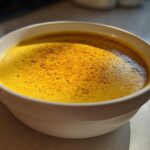 Close-up of a vibrant, creamy yellow cheese soup topped with dark spices in a white bowl.