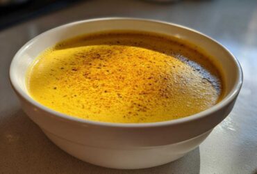 Close-up of a vibrant, creamy yellow cheese soup topped with dark spices in a white bowl.