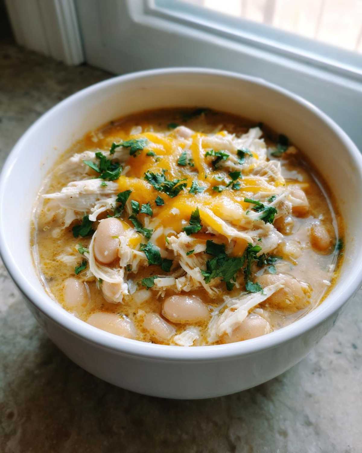 Close-up of a white bowl filled with creamy chicken chili, white beans, shredded chicken, melted cheese, and cilantro.