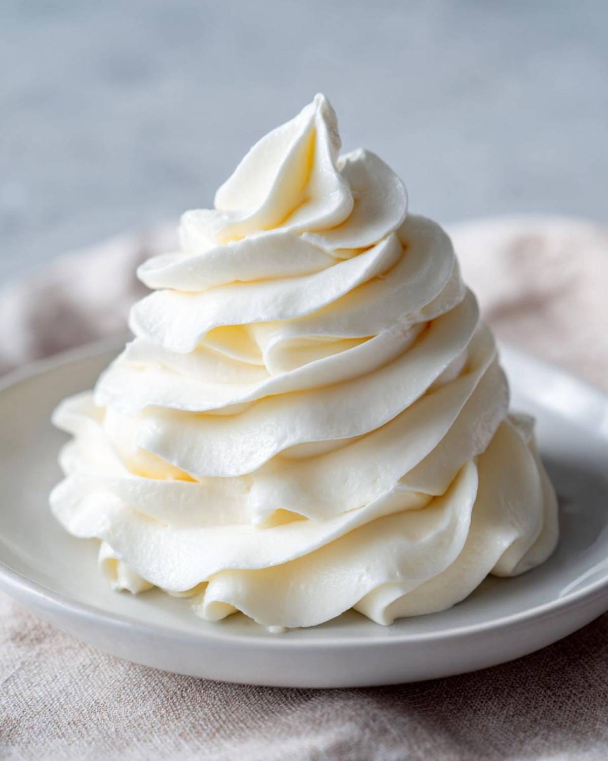 A tall, perfectly piped swirl of smooth, white cream cheese frosting on a small white plate.