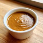 Close-up of a small white bowl filled with thick, rich brown peanut sauce, sitting on a wooden surface.