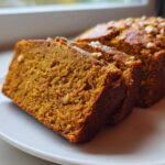 Close-up of moist, orange-hued pumpkin bread slices topped with nuts on a white plate.