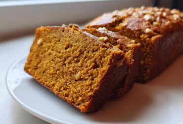 Close-up of moist, orange-hued pumpkin bread slices topped with nuts on a white plate.