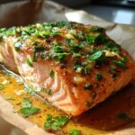 Close-up of a juicy, baked salmon fillet topped with fresh parsley and garlic glaze, resting on parchment paper.