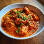 Close-up of rich, orange-red butter chicken pieces garnished with fresh cilantro in a white bowl.