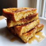Close-up of a tall stack of thick, golden brown french toast drizzled with syrup on a white plate.