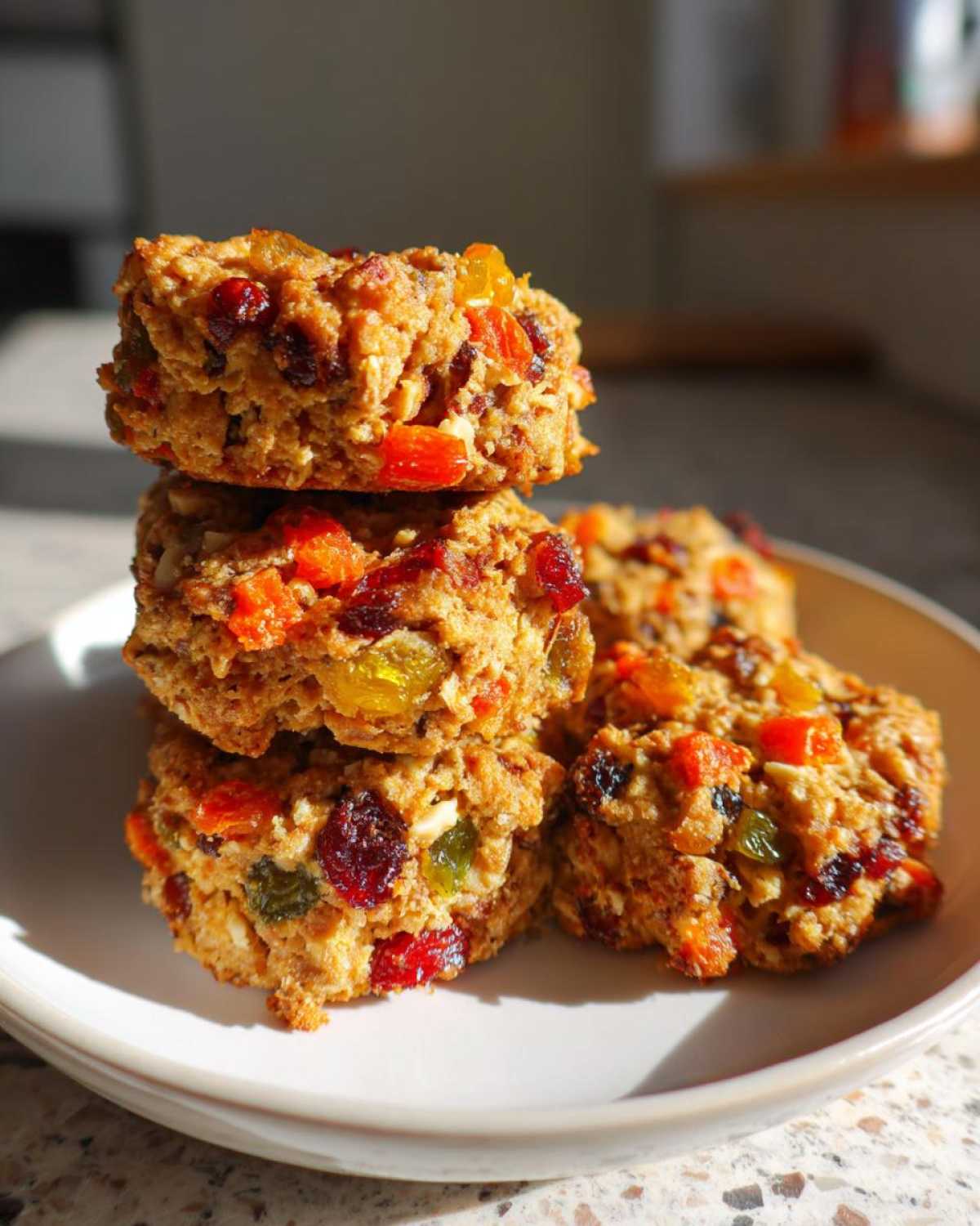A stack of three textured fruitcake cookies loaded with colorful candied fruit pieces, resting on a white plate.