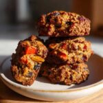 A stack of four rich, brown fruitcake cookies loaded with candied fruit pieces, sitting on a small plate.