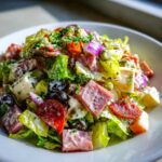 A close-up, vibrant bowl of grinder salad featuring chopped lettuce, cubed meats, cheese, red onion, and creamy dressing.
