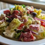 Close-up of a vibrant grinder salad featuring lettuce, cubed meats, feta cheese, red onion, and olives tossed in a creamy dressing.