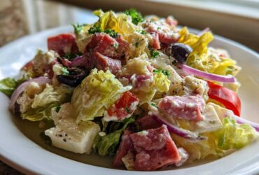 Close-up of a vibrant grinder salad featuring lettuce, cubed meats, feta cheese, red onion, and olives tossed in a creamy dressing.