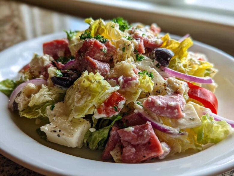 Close-up of a vibrant grinder salad featuring lettuce, cubed meats, feta cheese, red onion, and olives tossed in a creamy dressing.