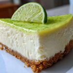 A close-up shot of a perfect slice of key lime cheesecake on a white plate, topped with lime glaze and a fresh lime slice.