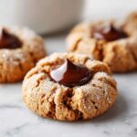A close-up of a perfectly baked, sugar-coated kiss cookie with a glossy dollop of melted chocolate in the center.