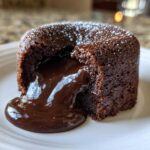 Close-up of a rich, dark chocolate lava cake with molten center oozing onto a white plate, dusted with powdered sugar.