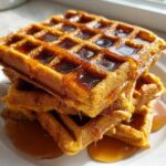 A stack of three golden brown pumpkin waffles generously drizzled with maple syrup.