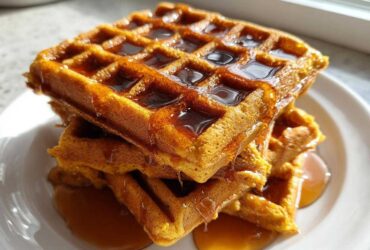 A stack of three golden brown pumpkin waffles generously drizzled with maple syrup.
