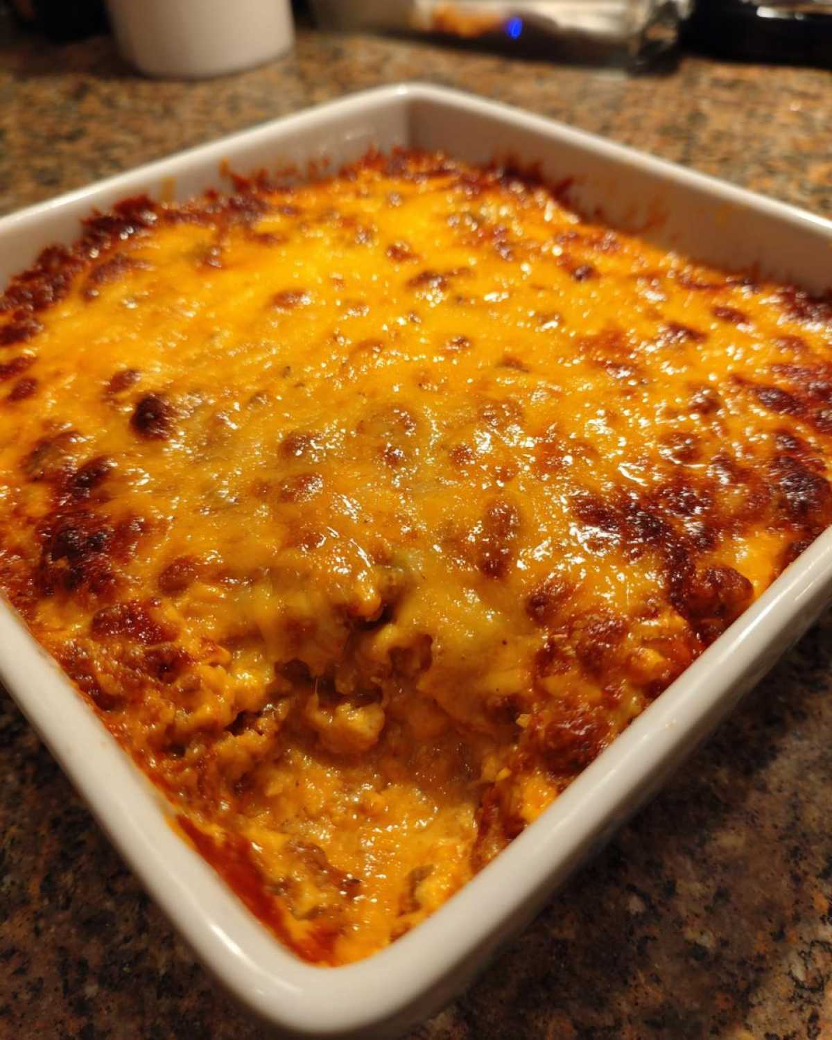 Close-up of a bubbly, baked chili cheese dip topped with melted, browned cheddar cheese in a square white baking dish.