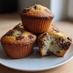 A stack of golden-brown banana chocolate chip muffins, one broken open to show the moist interior.