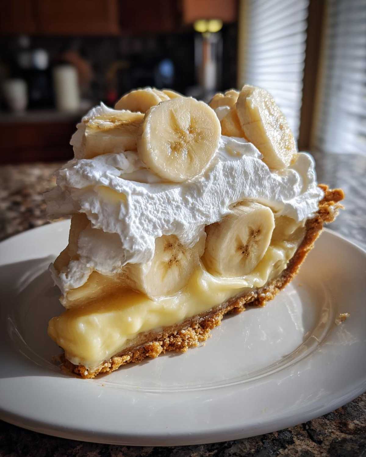 A tall slice of banana cream pie featuring a graham cracker crust, thick vanilla custard, whipped cream, and fresh banana slices.