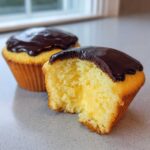 Close-up of a boston cream pie cupcake cut in half showing the vanilla cake, creamy filling, and chocolate glaze.