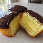 A boston cream pie cupcake cut in half showing the yellow cake, vanilla cream filling, and rich chocolate glaze.