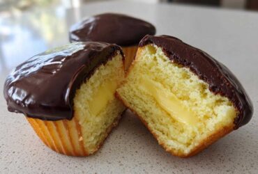 A boston cream pie cupcake cut in half showing the yellow cake, vanilla cream filling, and rich chocolate glaze.