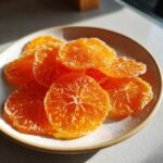 A close-up photo of several bright orange candied orange slices coated in sugar resting on a small ceramic plate.