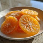 A small white bowl filled with glistening, sugar-coated candied orange slices, highlighted by bright sunlight.