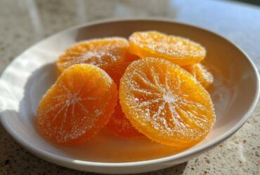 A small white bowl filled with glistening, sugar-coated candied orange slices, highlighted by bright sunlight.