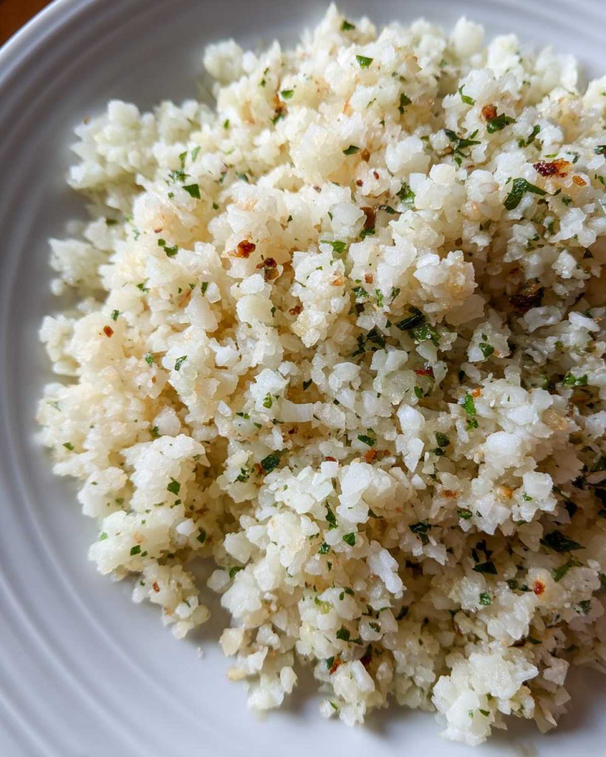 Close-up of fluffy, white cauliflower rice seasoned with green herbs and spices on a white plate.