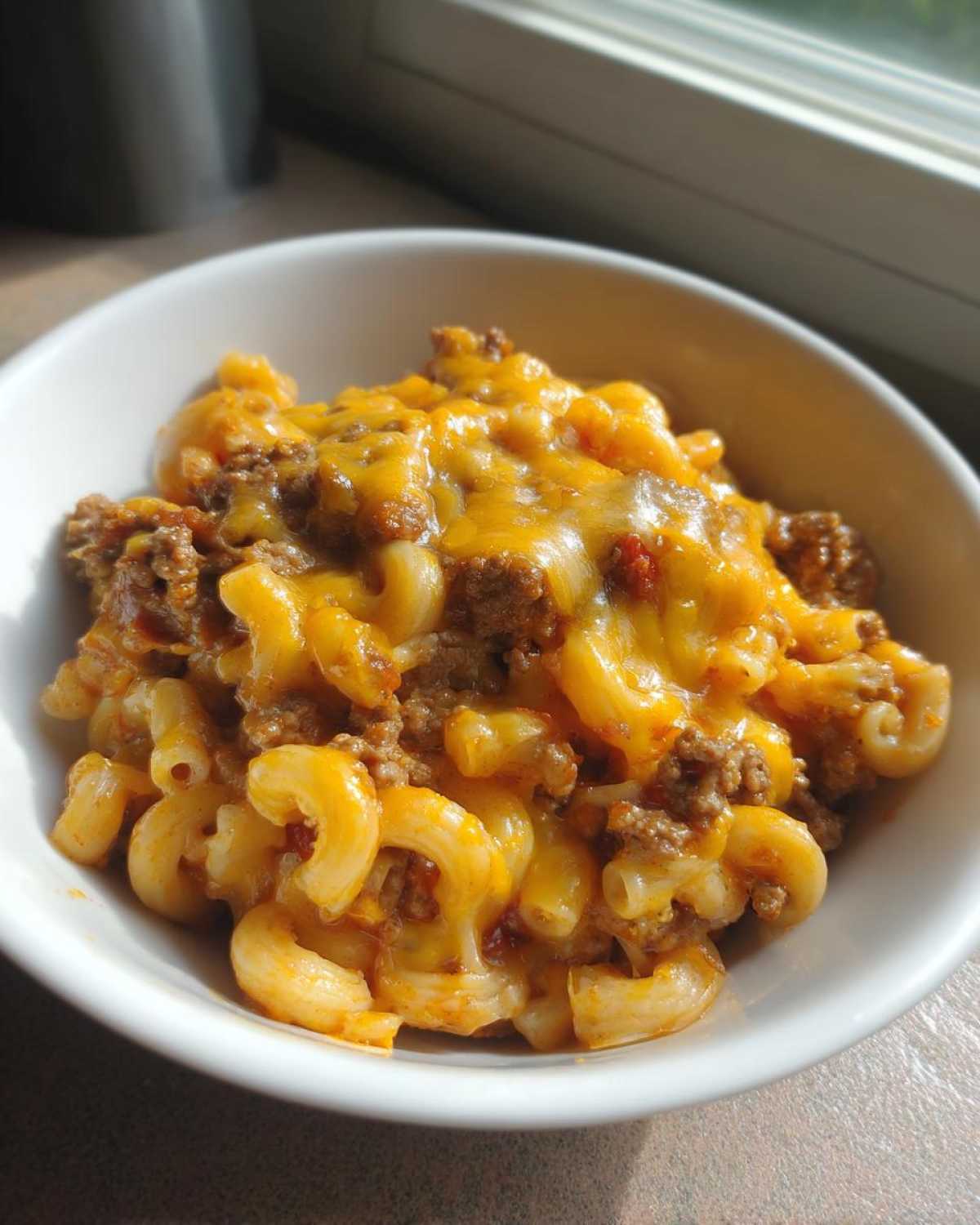 A close-up of a white bowl filled with cheesy cheeseburger casserole featuring elbow macaroni and ground beef.