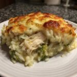 A close-up serving of creamy rice casserole with shredded chicken, broccoli, and a golden-brown melted cheese topping.