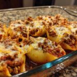 Close-up of baked stuffed shells topped with meat sauce and melted cheese in a clear glass baking dish.