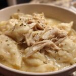 Close-up of a white bowl filled with creamy chicken and dumplings, topped with shredded chicken and herbs.
