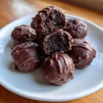A pile of rich, chocolate-covered Oreo balls on a white plate, one cut in half showing the dark, dense center.