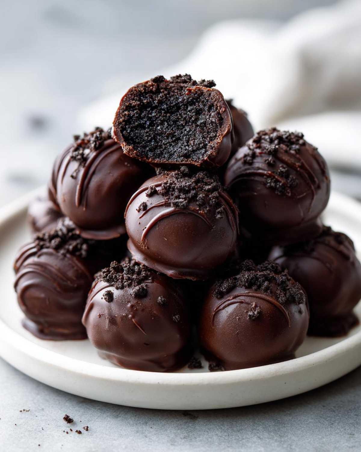 A stack of rich, chocolate-covered Oreo balls, one cut open to show the dark center, sprinkled with cookie crumbs.