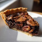 A close-up, sunlit slice of rich chocolate pecan pie showing the flaky crust and gooey filling loaded with pecans.