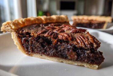 Close-up of a rich slice of chocolate pecan pie with a flaky crust and whole pecans on top.