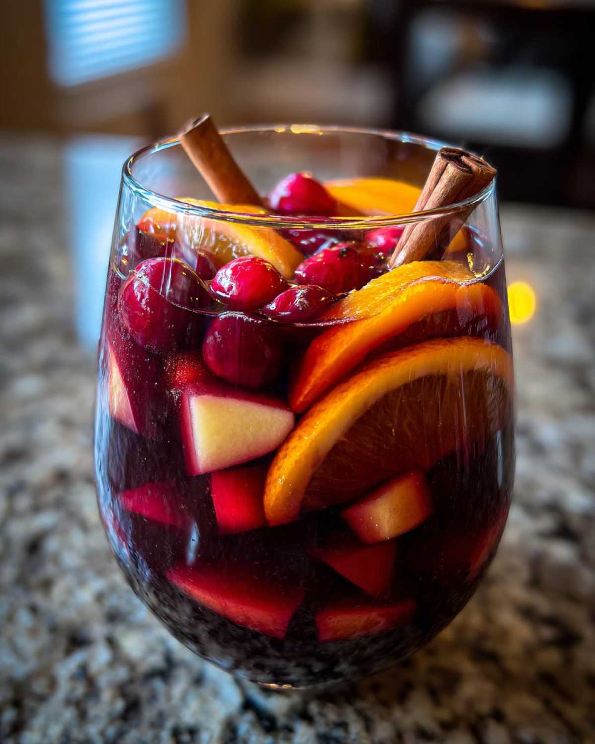 A close-up of a glass filled with rich red Christmas sangria, loaded with orange slices, cranberries, apples, and cinnamon sticks.