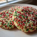Three round christmas sprinkle sugar cookies covered in red, green, and white nonpareils on a white plate.