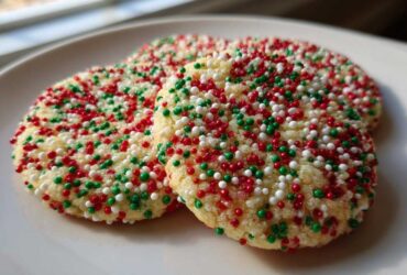 Three round christmas sprinkle sugar cookies covered in red, green, and white nonpareils on a white plate.