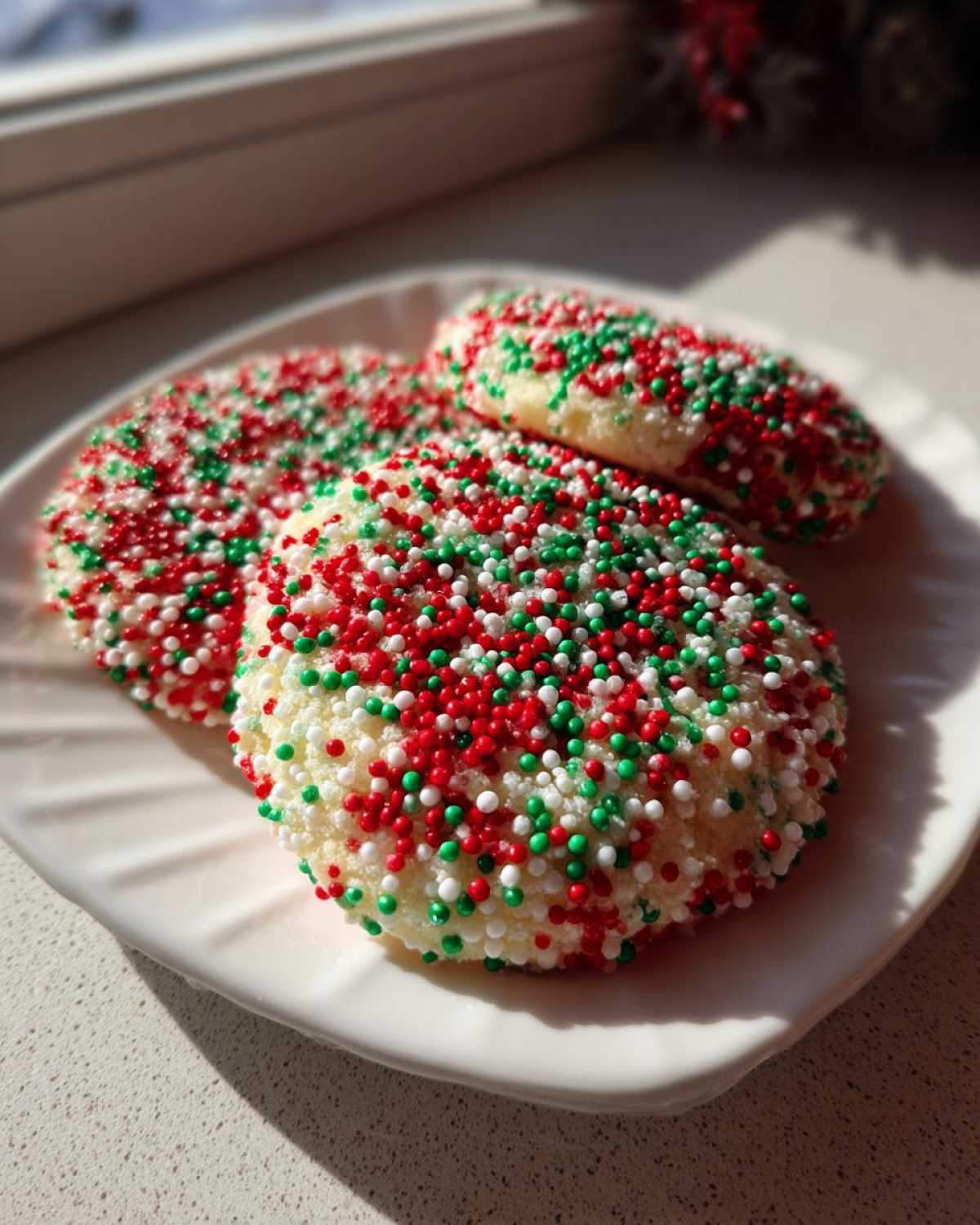 Three freshly baked christmas sprinkle sugar cookies coated heavily in red, green, and white nonpareils, resting on a white plate.