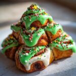 A stack of baked cinnamon rolls arranged in a Christmas tree shape, topped with bright green icing and festive sprinkles.