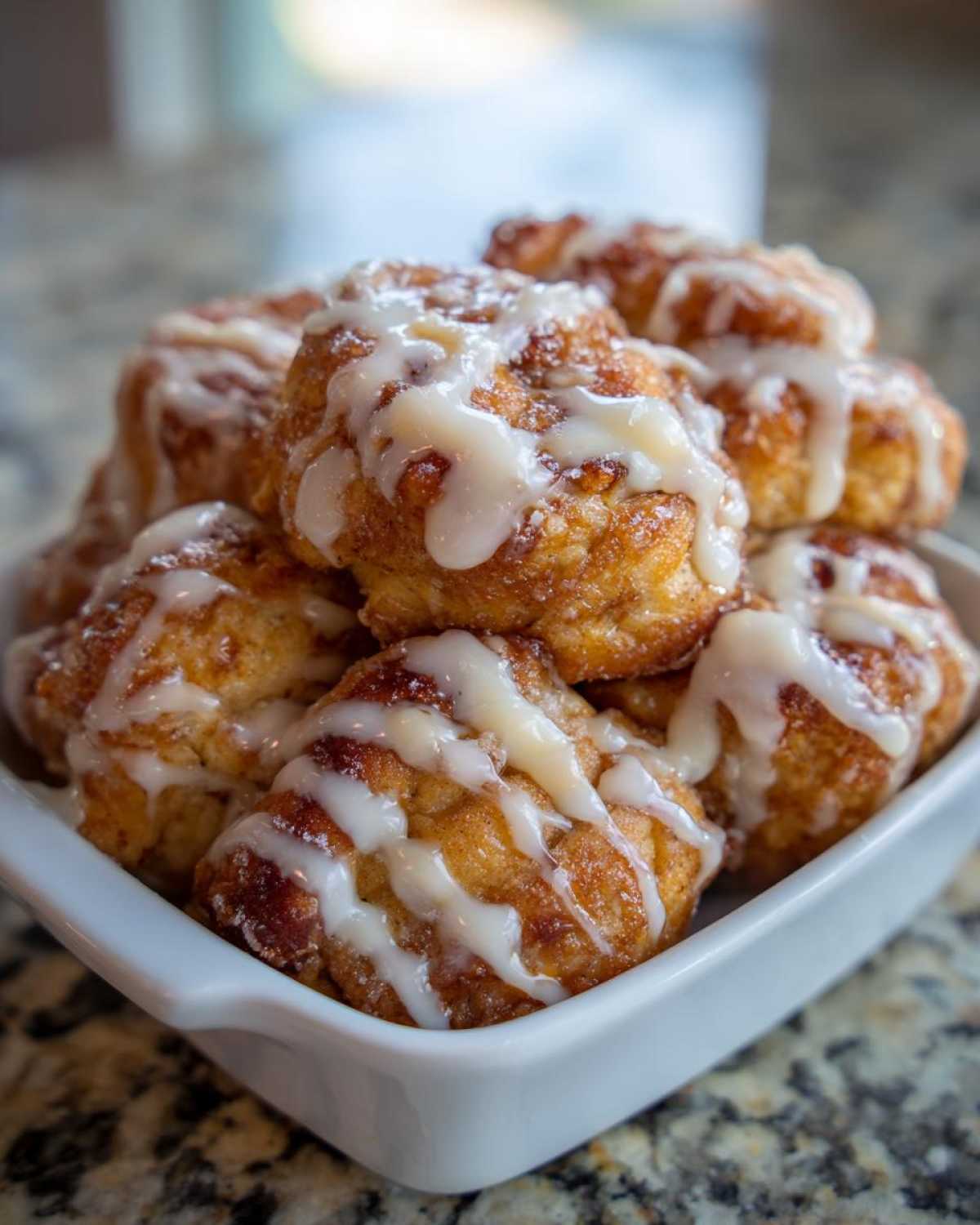A close-up of several golden brown cinnamon roll bites drizzled with white icing, piled high in a white square bowl.