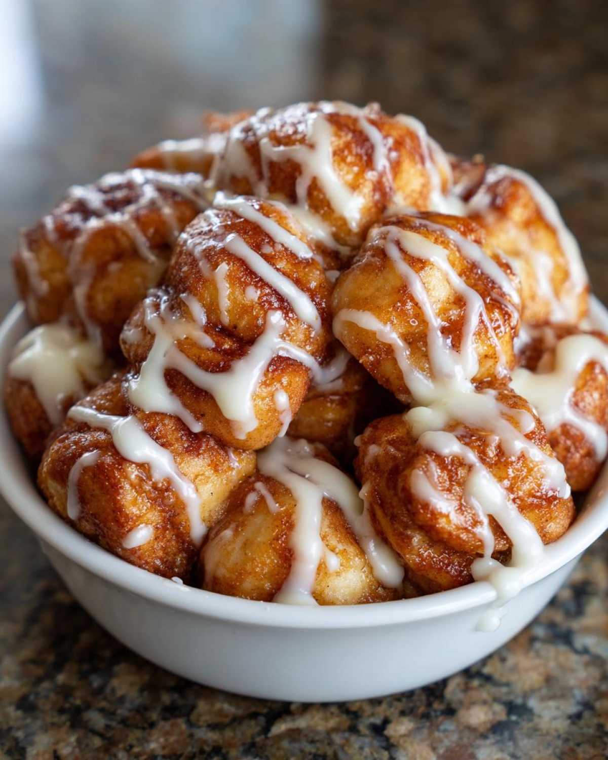 A white bowl overflowing with golden brown cinnamon roll bites drizzled generously with white icing.